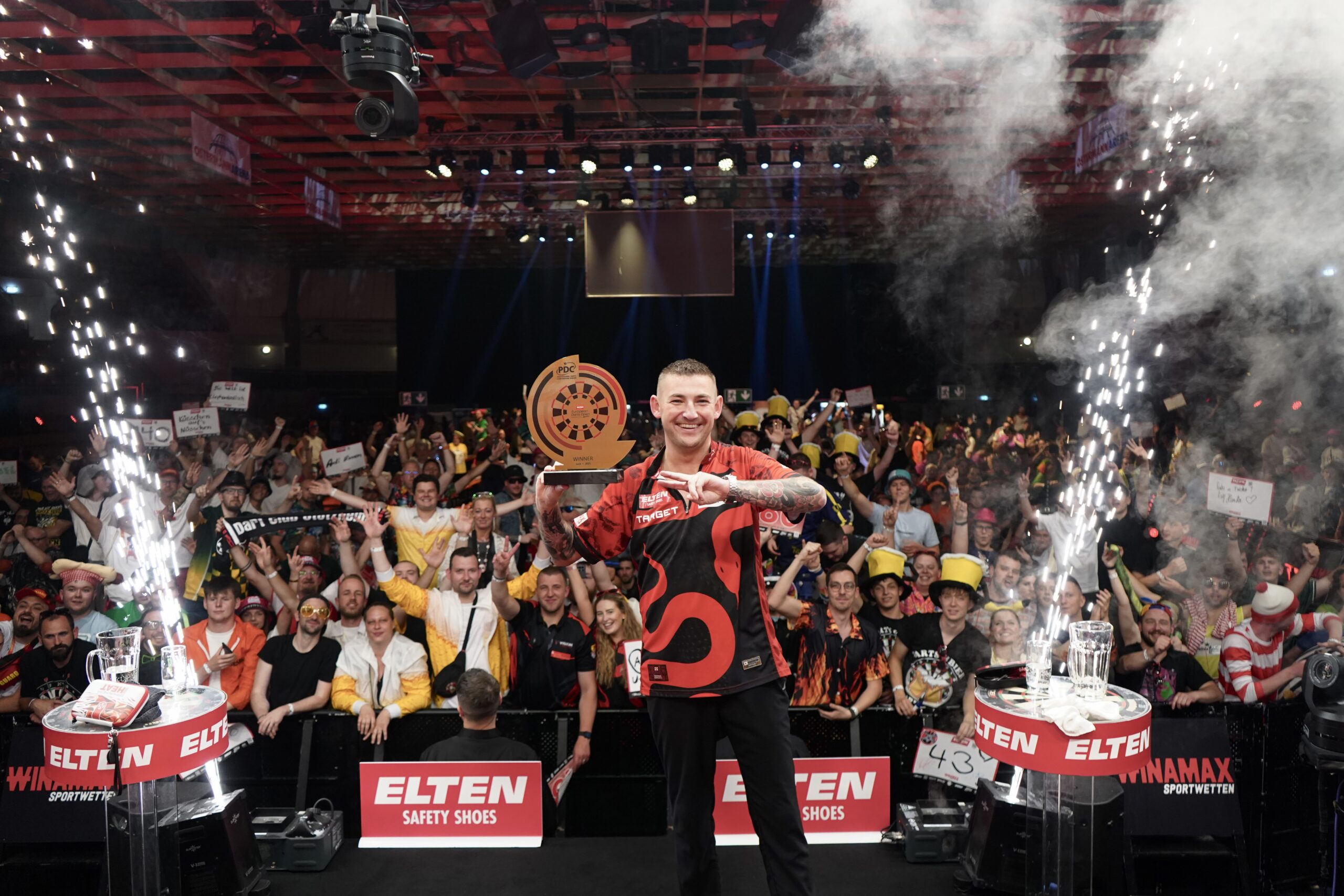Nathan Aspinall triumphiert mit Trophäe in der Hand vor jubelndem Publikum als Sieger der ELTEN Safety Shoes European Darts Open 2025.